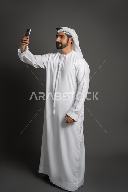 Using social media to communicate with friends, portrait of an Arab Gulf Emirati man wearing a kandura and ghutra making a video call using a mobile phone, full body image, black background