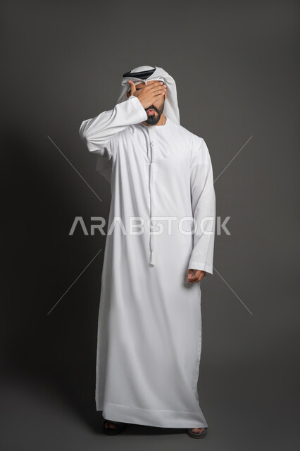 إيماءات وجه ويدين تدل على الندم والتحسر على موضوع معين، الانزعاج لدى سماع الأخبار السيئة، بورتريه لرجل عربي خليجي إماراتي يرتدي الكندورة والغترة البيضاء يضع يده على رأسه، صورة لكامل الجسم، خلفية سوداء