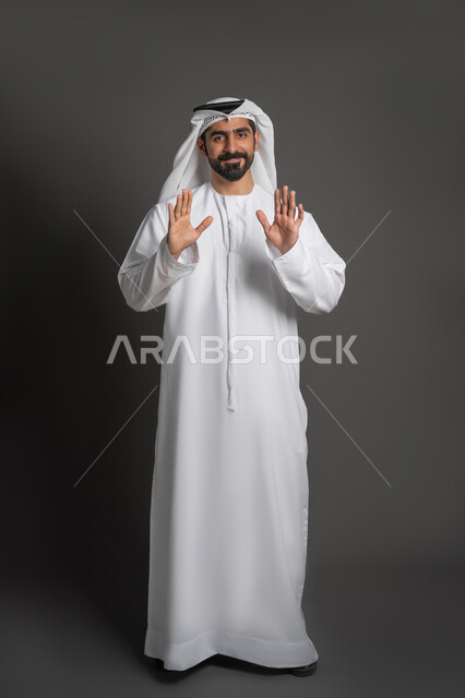 طلب المنع والتوقف عن فعل شيء ما، النظر إلى الكاميرا بتعابير البهجة، بورتريه لرجل عربي خليجي إماراتي يرتدي الكندورة والغترة يرفع يده بإيماءات الرفض والامتناع، صورة لكامل الجسم، خلفية سوداء