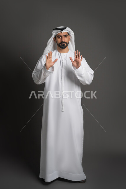 النظر إلى الكاميرا بتعابير الحزم والجدية، طلب المنع والتوقف عن فعل شيء ما، بورتريه لرجل عربي خليجي إماراتي يرتدي الكندورة والغترة يرفع يده بإيماءات الرفض والامتناع، صورة لكامل الجسم، خلفية سوداء