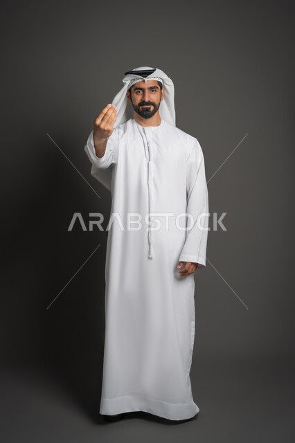 الهدوء وطلب الإنتظار، بورتريه لرجل عربي خليجي إماراتي يرتدي الكندورة والغترة البيضاء يرفع يده بإيماءات الصبر والتأني، إمهال الوقت للقيام بشيء ما، صورة لكامل الجسم، خلفية سوداء