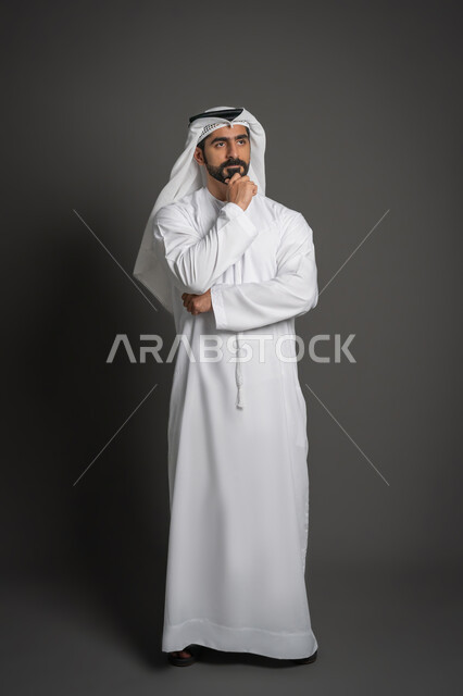 التخطيط للمستقبل ووضع الأهداف، النظر إلى شيء ما بتعابير التركيز، بورتريه لرجل عربي خليجي إماراتي يرتدي الكندورة والغترة البيضاء يضع يده على ذقنه بإيماءات التفكير, صورة لكامل الجسم، خلفية سوداء
