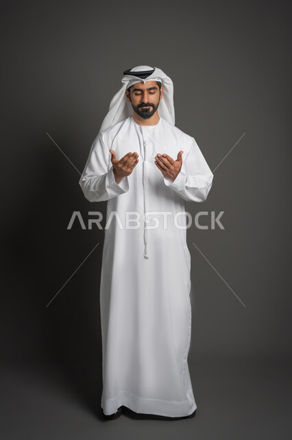 تعابير الخشوع والتضرع إلى الله، مفهوم الأناقة والاهتمام بالمظهر الخارجي, بورتريه لرجل عربي خليجي إماراتي يرتدي الكندورة البيضاء والغترة يقوم برفع يديه إلى الأعلى بإيماءات الدعاء, صورة لكامل الجسم، خلفية سوداء