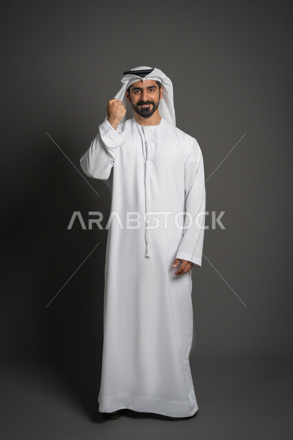 مفهوم الأناقة والرجولة والاهتمام بالمظهر الخارجي, بورتريه لرجل عربي خليجي إماراتي يرتدي الكندورة البيضاء والغترة يرفع يده بتعابير القوة والتحدي, الوقوف باستقامة والنظر إلى الكاميرا بايماءات الثقة بالنفس, صورة لكامل الجسم، خلفية سوداء