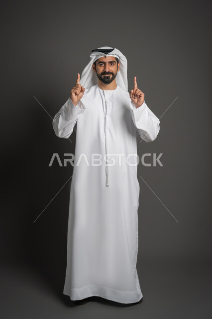 رفع إصبعي السبابة بإيماءات النطق بالشهادتين، بورتريه لرجل عربي خليجي إماراتي يرتدي الكندورة والغترة ينظر إلى الأعلى بتعابير الدعاء والتضرع إلى الله، صورة لكامل الجسم، خلفية سوداء
