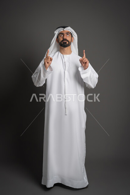 النظر إلى الأعلى بتعابير الدعاء والتضرع إلى الله، بورتريه لرجل عربي خليجي إماراتي يرتدي الكندورة والغترة يرفع إصبعي السبابة بإيماءات النطق بالشهادتين، صورة لكامل الجسم، خلفية سوداء