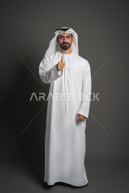 مفهوم الأناقة والرجولة والاهتمام بالمظهر الخارجي, بورتريه لرجل عربي خليجي إماراتي يرتدي الكندورة البيضاء والغترة ويرفع اصبع الابهام بإيماءة إلى الموافقة والقبول, الوقوف باستقامة والنظر إلى الكاميرا بتعابير الثقة بالنفس, صورة لكامل الجسم، خلفية سوداء