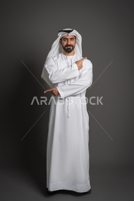 Standing straight and looking at the camera with a confident expression, portrait of an Emirati Gulf Arab man wearing a white kandura and ghutra pointing in two different directions using the index fingers of both hands, taking care of one's appearance, full body photo, black background