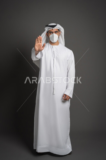 تجنب انتشار العدوى بكوفيد 19، منع الاقتراب للحفاظ على السلامة, اتباع الإجراءات الاحترازية للوقاية من فايروس كورونا، بورتريه لرجل عربي إماراتي خليجي يرتدي الكندورة والغترة يضع الكمامة يرفع كف يده لأعلى بتعابير التحذير والتوقف, صورة لكامل الجسم، خلفية سوداء