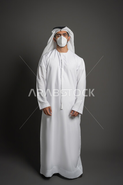 إتباع الإجراءات الوقائيه ضد الفايروسات، الوقوف باستقامة والنظر غلى الكاميرا بتعابير الحزم والجدية، بورتريه لرجل عربي خليجي اماراتي يرتدي الكندوره والغتره يضع القناع الطبي الوقائي للوقايه من فايروس كورونا، صورة لكامل الجسم، خلفيه سوداء