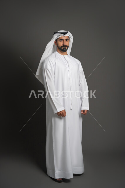 مفهوم الرجولة والثقة بالنفس، التقاط الصور الشخصية للوثائق الرسمية، بورتريه لرجل عربي خليجي إماراتي يرتدي الكندورة والغترة ينظر الى الكاميرا بتعابير الحزم والجدية، الوقوف باستعداد واستقامة، الأناقة والاهتمام بالمظهر الخارجي، صورة الجسم بالكامل، خلفية سوداء