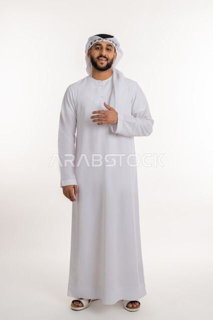 Feeling reassured and comfortable, portrait of a smiling Emirati Gulf Arab man wearing a kandora and ghutra, looking at the camera, placing his hand on his chest with greeting and welcome gestures, expressions of pleasure and joy, full-length body portrait, white background