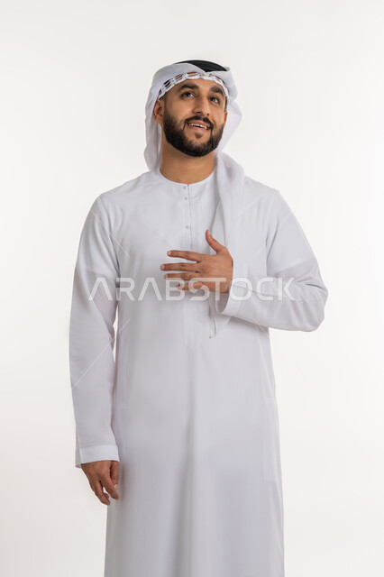 الشعور بالاطمئنان والراحة، بورتريه لرجل عربي خليجي إماراتي مبتسم يرتدي الكندورة والغترة ينظر إلى شيء ما يضع يده على صدره بإيماءات الاستقبال والترحيب, تعابير السرور والفرح، خلفية بيضاء