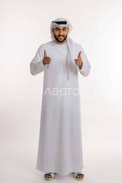 تحديد موقع معين، بورتريه لرجل عربي خليجي إماراتي مبتسم يرتدي الكندورة والغترة ينظر الى الكاميرا ييرفع يده بإيماءات الإشارة إلى شيء ما، تعابير الفرح والسرور, صورة الجسم بالطول الكامل، خلفية بيضاء