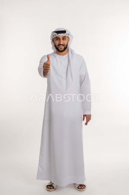 تعابير القبول والفرح والسرور، الاهتمام بالأناقة والمظهر الخارجي، بورتريه لرجل عربي خليجي إماراتي يرتدي الكندورة والغترة ينظر الى الكاميرا يرفع إصبع السبابة للاعلى بإيماءات الإعجاب والرضا، صورة الجسم بالطول الكامل، خلفية بيضاء