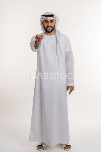 الاهتمام بالأناقة والمظهر الخارجي، تعابير السرور والثقة بالنفس، بورتريه لرجل عربي خليجي إماراتي يرتدي الكندورة والغترة ينظر الى الكاميرا ييشير بإصبع السبابة، صورة الجسم بالطول الكامل، خلفية بيضاء