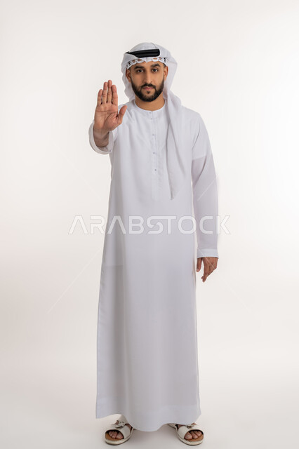 مفهوم الرفض والامتناع والانزعاج، النهي عن فعل أمر ما، بورتريه لرجل عربي خليجي إماراتي يرتدي الكندورة والغترة ينظر الى الكاميرا يرفع يده بإيماءات التوقف وعدم الاقتراب، صورة الجسم بالطول الكامل، خلفية بيضاء
