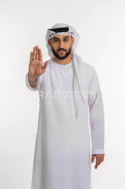 مفهوم الرفض والامتناع والانزعاج، النهي عن فعل أمر ما، بورتريه لرجل عربي خليجي إماراتي يرتدي الكندورة والغترة ينظر الى الكاميرا يرفع يده بإيماءات التوقف وعدم الاقتراب، خلفية بيضاء