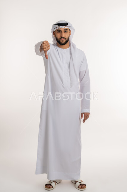 مفهوم التقييم السلبي والانزعاج، الفشل في تحقيق أمر ما، بورتريه لرجل عربي خليجي إماراتي يرتدي الكندورة والغترة ينظر الى الكاميرا يشير بإصبع الابهام الى الاسفل بإيماءات الرفض وعدم القبول والرضا، صورة الجسم بالطول الكامل، خلفية بيضاء