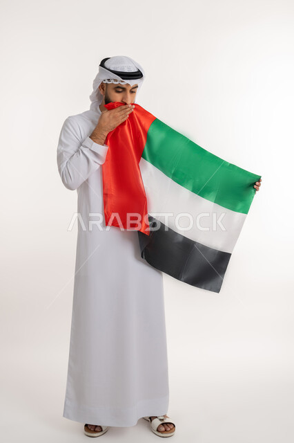 الاحتفال باليوم الوطني 2 ديسمبر، مفهوم حب الوطن والانتماء إليه، إحياء ذكرى استقلال البلاد، بورتريه لرجل عربي خليجي إماراتي يرتدي الكندورة والغترة يقوم بتقبيل علم الإمارات المتحدة، إيماءات الاعتزاز والفخر، صورة الجسم بالطول الكامل، خلفية بيضاء