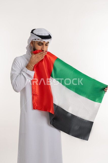 الاحتفال باليوم الوطني 2 ديسمبر، مفهوم حب الوطن والانتماء إليه، إحياء ذكرى استقلال البلاد، بورتريه لرجل عربي خليجي إماراتي يرتدي الكندورة والغترة يقوم بتقبيل علم الإمارات المتحدة، إيماءات الاعتزاز والفخر، خلفية بيضاء