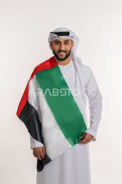 مفهوم حب الوطن والانتماء إليه، إحياء ذكرى استقلال البلاد، بورتريه لرجل عربي خليجي إماراتي يرتدي الكندورة والغترة ينظر الى الكاميرا يضع علم الإمارات المتحدة على كتفه، الاحتفال باليوم الوطني 2 ديسمبر، إيماءات الاعتزاز والفخر والثقة بالنفس، خلفية بيضاء