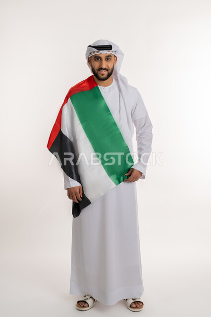 الاحتفال باليوم الوطني 2 ديسمبر، مفهوم حب الوطن والانتماء إليه، إحياء ذكرى استقلال البلاد، بورتريه لرجل عربي خليجي إماراتي يرتدي الكندورة والغترة ينظر الى الكاميرا يضع علم الإمارات على كتفه، إيماءات الاعتزاز والفخر، صورة الجسم بالطول الكامل، خلفية بيضاء