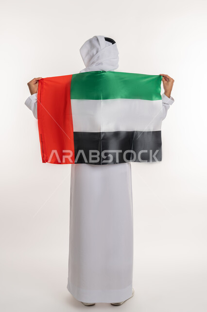 مفهوم حب الوطن والانتماء إليه، إحياء ذكرى استقلال البلاد، بورتريه من الخلف لرجل عربي خليجي إماراتي يرتدي الكندورة والغترة يضع علم الإمارات المتحدة على كتفيه، الاحتفال باليوم الوطني 2 ديسمبر، صورة الجسم بالطول الكامل، خلفية بيضاء