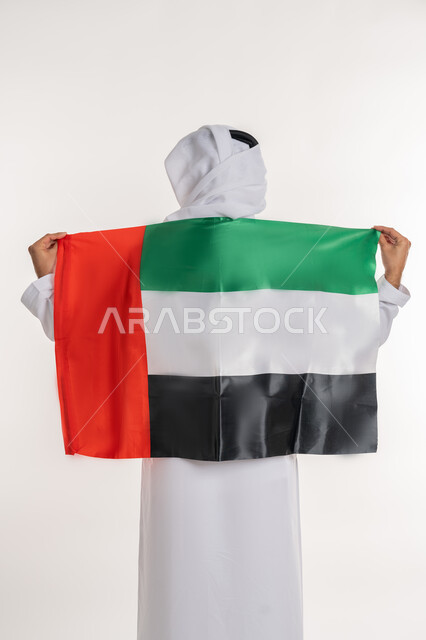 مفهوم حب الوطن والانتماء إليه، إحياء ذكرى استقلال البلاد، بورتريه من الخلف لرجل عربي خليجي إماراتي يرتدي الكندورة والغترة يضع علم الإمارات المتحدة على كتفيه، الاحتفال باليوم الوطني 2 ديسمبر، خلفية بيضاء