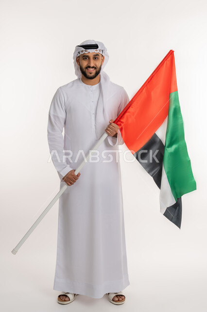 Commemorating the country's independence, the concept of love for the homeland and belonging to it, portrait of an Emirati Gulf Arab man wearing a kandora and ghutra looking at the camera raising the UAE flag, celebrating National Day December 2, gestures of pride and honor, full-length body portrait, white background