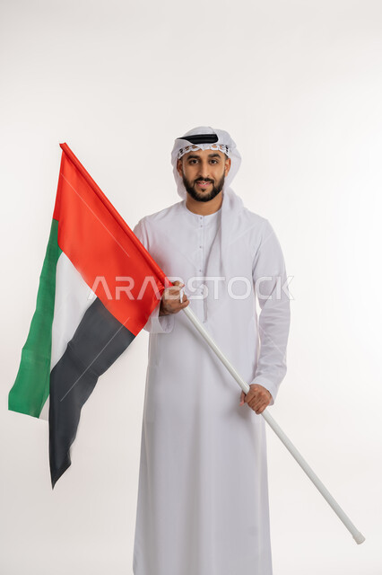 إحياء ذكرى استقلال البلاد، مفهوم حب الوطن والانتماء إليه، بورتريه لرجل عربي خليجي إماراتي يرتدي الكندورة والغترة ينظر الى الكاميرا يرفع علم الإمارات المتحدة، الاحتفال باليوم الوطني 2 ديسمبر، إيماءات الاعتزاز والفخر والثقة بالنفس، خلفية بيضاء