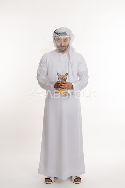 استخدام خشب العود في المناسبات والأعياد, التطيب بالروائح الفريدة، بورتريه لرجل عربي خليجي اماراتي يرتدي الكندورة والغترة يتعطر بحامل البخور ( المبخرة ), أصالة العادات والتقاليد، الاهتمام بالأناقة والمظهر الخارجي، صورة الجسم بالطول الكامل، خلفية بيضاء