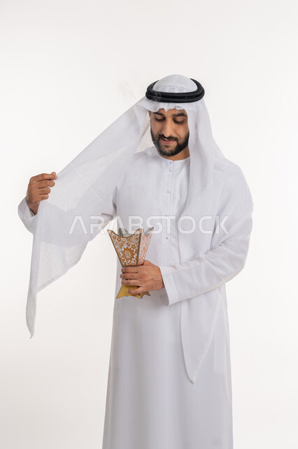 الاهتمام بالأناقة والمظهر الخارجي، استخدام خشب العود في المناسبات والأعياد, التطيب بالروائح الفريدة، بورتريه لرجل عربي خليجي اماراتي يرتدي الكندورة والغترة يقوم بتعطير ملابسه بحامل البخور ( المبخرة ), أصالة العادات والتقاليد، خلفية بيضاء