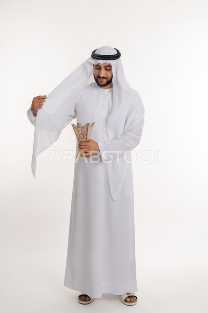 مفهوم الأناقة والرجولة، استخدام خشب العود في المناسبات والأعياد, التطيب بالروائح الفريدة، بورتريه لرجل عربي خليجي اماراتي يرتدي الكندورة والغترة يقوم بتعطير ملابسه بحامل البخور ( المبخرة ), أصالة العادات والتقاليد، صورة الجسم بالطول الكامل، خلفية بيضاء