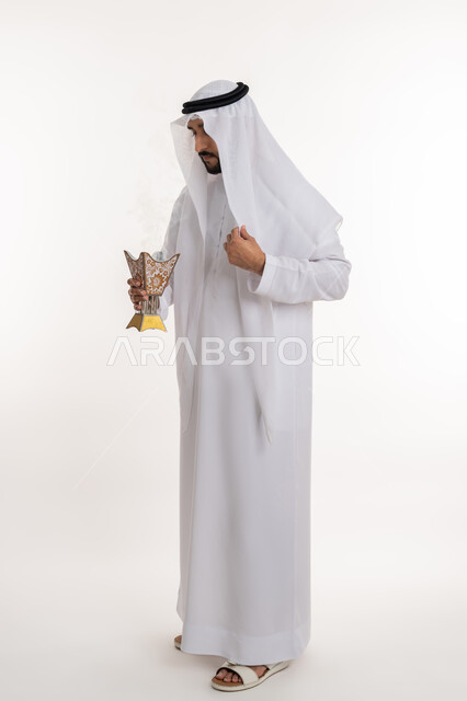 مفهوم الأناقة والرجولة، استخدام خشب العود في المناسبات والأعياد, التطيب بالروائح الفريدة، بورتريه لرجل عربي خليجي اماراتي يرتدي الكندورة والغترة يقوم بتعطير ملابسه بحامل البخور ( المبخرة ), أصالة العادات والتقاليد، صورة الجسم بالطول الكامل، خلفية بيضاء