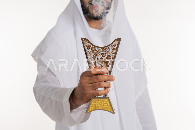 أصالة العادات والتقاليد، استخدام خشب العود في المناسبات والأعياد, التطيب بالروائح الفريدة، بورتريه مقرب لرجل عربي خليجي اماراتي يرتدي الكندورة والغترة يتعطر بحامل البخور ( المبخرة ), خلفية بيضاء