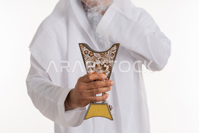 أصالة العادات والتقاليد، استخدام خشب العود في المناسبات والأعياد, التطيب بالروائح الفريدة، بورتريه مقرب لرجل عربي خليجي اماراتي يرتدي الكندورة والغترة يتعطر بحامل البخور ( المبخرة ), خلفية بيضاء