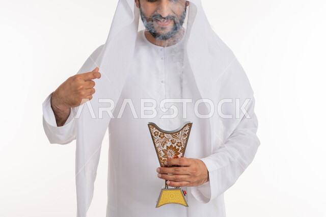 أصالة العادات والتقاليد، استخدام خشب العود في المناسبات والأعياد, التطيب بالروائح الفريدة، بورتريه مقرب لرجل عربي خليجي اماراتي يرتدي الكندورة والغترة يتعطر بحامل البخور ( المبخرة ), خلفية بيضاء