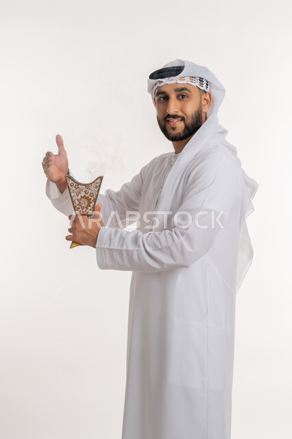 التطيب بالروائح الفريدة، استخدام خشب العود في المناسبات والأعياد, بورتريه لرجل عربي خليجي اماراتي مبتسم يرتدي الكندورة والغترة ينظر الى الكاميرا يتعطر بحامل البخور ( المبخرة ), أصالة العادات والتقاليد، الاهتمام بالأناقة والمظهر الخارجي، خلفية بيضاء