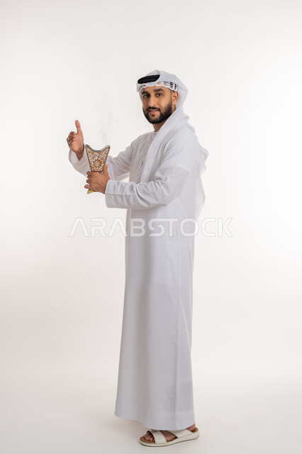 استخدام خشب العود في المناسبات والأعياد, تعابير السرور والفرح، التطيب بالروائح الفريدة، بورتريه لرجل عربي خليجي اماراتي مبتسم يرتدي الكندورة والغترة ينظر الى الكاميرا يتعطر بحامل البخور( المبخرة ), أصالة العادات والتقاليد، صورة الجسم بالطول الكامل، خلفية بيضاء