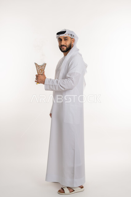 The use of agarwood on occasions and holidays, expressions of happiness and joy, wearing unique scents, a portrait of a smiling Arab Gulf Emirati man wearing a kandora and ghutra, looking at the camera, perfumed with an incense holder (incense burner), authenticity of customs and traditions, full-length body portrait, white background