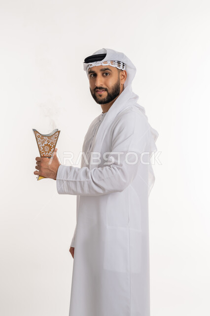 التطيب بالروائح الفريدة، استخدام خشب العود في المناسبات والأعياد, بورتريه لرجل عربي خليجي اماراتي مبتسم يرتدي الكندورة والغترة ينظر الى الكاميرا يتعطر بحامل البخور ( المبخرة ), أصالة العادات والتقاليد، الاهتمام بالأناقة والمظهر الخارجي، خلفية بيضاء