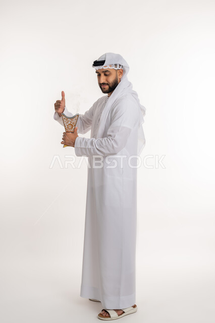 التطيب بالروائح الفريدة، استخدام خشب العود في المناسبات والأعياد, بورتريه لرجل عربي خليجي اماراتي مبتسم يرتدي الكندورة والغترة يتعطر بحامل البخور ( المبخرة ), أصالة العادات والتقاليد، الاهتمام بالأناقة والمظهر الخارجي، صورة الجسم بالطول الكامل، خلفية بيضاء