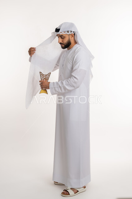 استخدام خشب العود في المناسبات والأعياد, التطيب بالروائح الفريدة، بورتريه لرجل عربي خليجي اماراتي يرتدي الكندورة والغترة يقوم بتعطير ملابسه  بحامل البخور ( المبخرة ), أصالة العادات والتقاليد، الاهتمام بالمظهر الخارجي، صورة الجسم بالطول الكامل، خلفية بيضاء