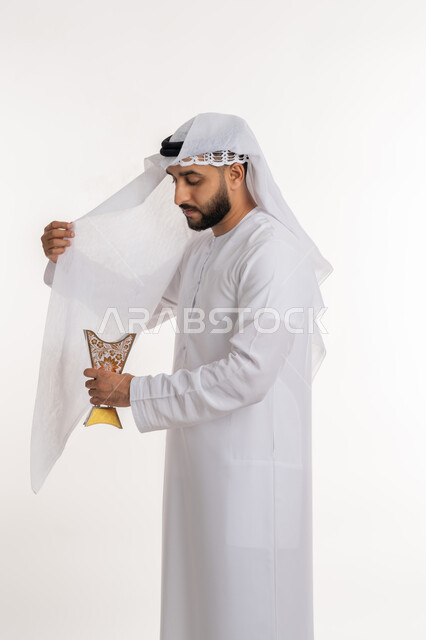 Paying attention to elegance and external appearance, using agarwood on special occasions and holidays, perfuming with unique scents, a portrait of an Arab Gulf Emirati man wearing a kandora and ghutra, perfuming his clothes with an incense holder (incense burner), authenticity of customs and traditions, white background