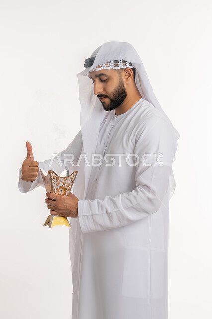 الاهتمام بالأناقة والمظهر الخارجي، استخدام خشب العود في المناسبات والأعياد, التطيب بالروائح الفريدة، بورتريه لرجل عربي خليجي اماراتي يرتدي الكندورة والغترة يقوم بتعطير ملابسه  بحامل البخور ( المبخرة ), أصالة العادات والتقاليد، خلفية بيضاء