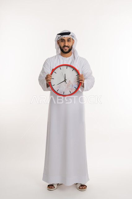 التنبيهات والتذكيرات الثابته، أهمية الدقة والالتزام في المواعيد، بورتريه لرجل عربي خليجي اماراتي مبتسم يرتدي الكندورة والغترة ينظر الى الكاميرا يحمل بيده ساعة رقمية كبيرة, إنجاز المهامات في الوقت، صورة الجسم الطول الكامل, خلفية بيضاء