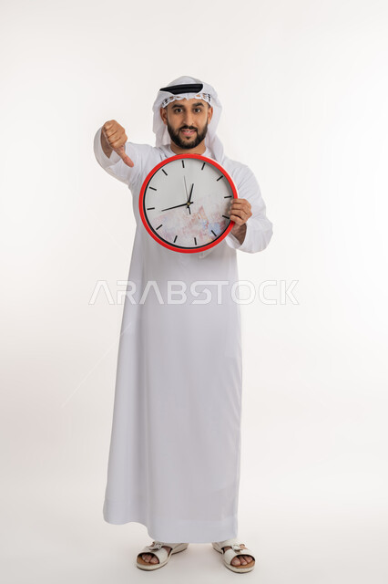 التأخير وعدم الالتزام بالمواعيد، التنبيهات والتذكيرات الثابته، بورتريه لرجل عربي خليجي اماراتي يرتدي الكندورة والغترة ينظر الى الكاميرا يحمل بيده ساعة رقمية كبيرة يرفع إصبع السبابة الى الاسفل بإيماءات عدم الرضا, صورة الجسم الطول الكامل, خلفية بيضاء
