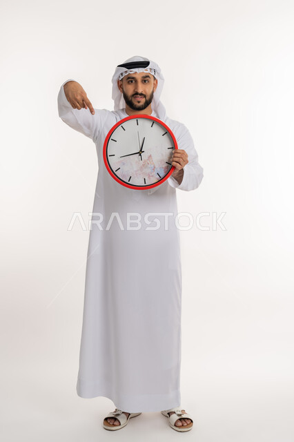 التنبيهات والتذكيرات الثابته، الدقة في المواعيد، بورتريه لرجل عربي خليجي اماراتي يرتدي الكندورة والغترة ينظر الى الكاميرا يحمل بيده ساعة رقمية كبيرة يشير بإصبع السبابة الى التوقيت, إنجاز المهامات في الوقت، صورة الجسم الطول الكامل, خلفية بيضاء