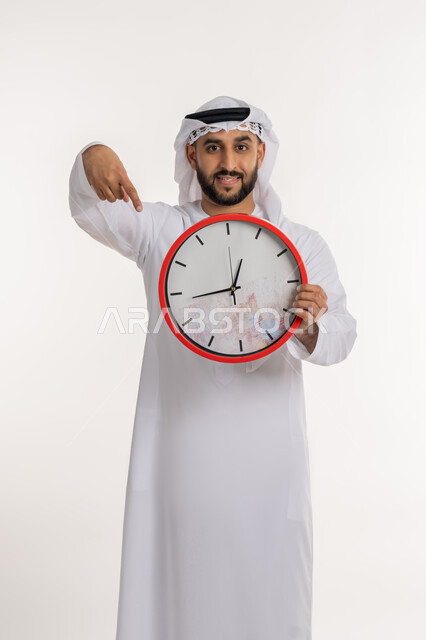 التنبيهات والتذكيرات الثابته، الدقة في المواعيد، بورتريه لرجل عربي خليجي اماراتي يرتدي الكندورة والغترة ينظر الى الكاميرا يحمل بيده ساعة رقمية كبيرة يشير بإصبع السبابة الى التوقيت, إنجاز المهامات في الوقت، خلفية بيضاء
